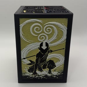 May include: Black rectangular box featuring a silhouette of a character in a kneeling pose with a staff, set against a yellow-green background with white swirling designs. The box has a decorative top with circular patterns.