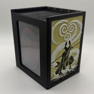 May include: Black card storage box with a decorative panel featuring a character silhouette and swirling white and gold accents. The box has a clear window to display a card. The box is designed to hold trading cards.