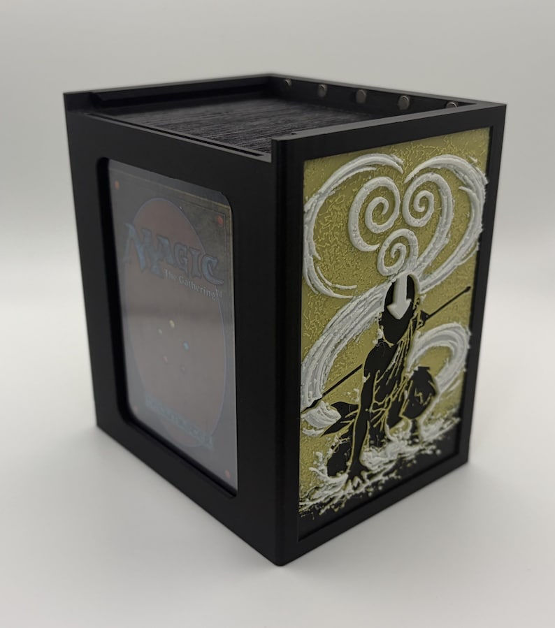 May include: Black card storage box with a decorative panel featuring a character silhouette and swirling white and gold accents. The box has a clear window to display a card. The box is designed to hold trading cards.