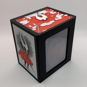 Puede incluir: Caja negra para guardar cartas con una tapa roja con un diseño blanco y plateado. Los lados muestran un personaje en rojo y negro. La caja tiene una ventana transparente para ver las cartas dentro.