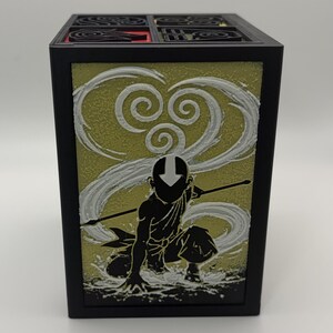 May include: Black square box featuring a silhouette of a character with a staff, set against a yellow and white swirling background. The top of the box has a black grid design with red accents. The box is likely a decorative item or storage container.