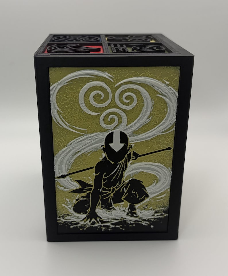 May include: Black square box featuring a silhouette of a character with a staff, set against a yellow and white swirling background. The top of the box has a black grid design with red accents. The box is likely a decorative item or storage container.