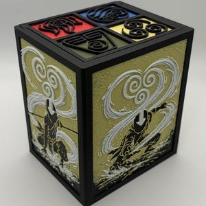 May include: A black, cube-shaped box with detailed artwork. The top features four colored squares with symbols. The sides display a golden background with white and black figures in action poses. The box has a black frame.