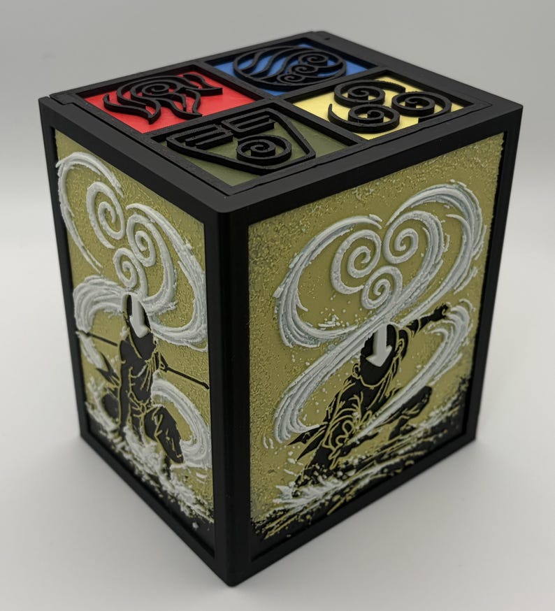 May include: A black, cube-shaped box with detailed artwork. The top features four colored squares with symbols. The sides display a golden background with white and black figures in action poses. The box has a black frame.