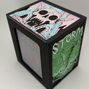 May include: A black rectangular card storage box featuring a top panel with a skull illustration in pastel pink and blue. The side panel displays a green graphic of a tornado with the word STORM printed in white, and a Magic card is visible inside.