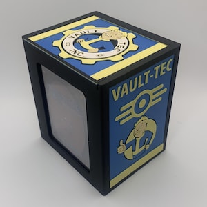 May include: Black box featuring Vault-Tec branding in blue and yellow. The design includes the Vault-Tec logo and a cartoon character. The box has a clear window on one side, and is likely a display case or storage unit.