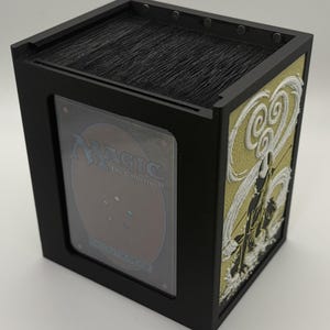 May include: Black card storage box with a clear window displaying a Magic: The Gathering card. The side panel features a gold and white relief design. The box is filled with black cards.