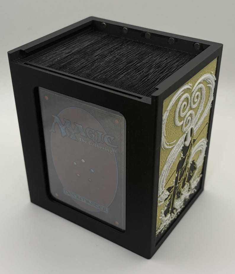 May include: Black card storage box with a clear window displaying a Magic: The Gathering card. The side panel features a gold and white relief design. The box is filled with black cards.