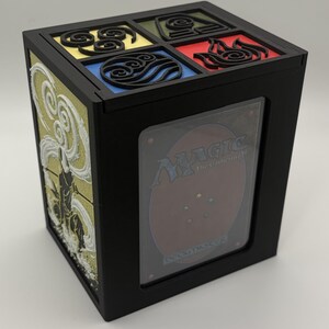 May include: Black card storage box with a clear window displaying a Magic: The Gathering card. The top features four colored squares with elemental symbols. The side has a white and gold relief design.
