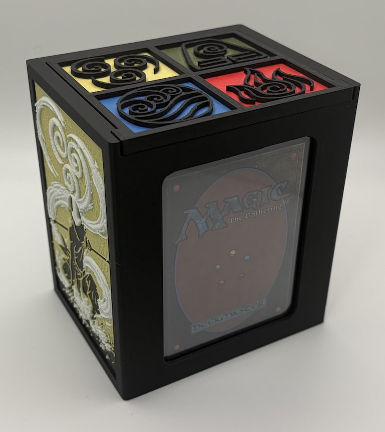 May include: Black card storage box with a clear window displaying a Magic: The Gathering card. The top features four colored squares with elemental symbols. The side has a white and gold relief design.