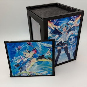 Könnte beinhalten: Eine schwarze, rechteckige Aufbewahrungsbox mit Magnetpaneelen, die einen blauhaarigen Anime-Charakter in einer Konzertumgebung zeigen. Ein passendes quadratisches Paneel mit demselben Charakter steht davor und zeigt leuchtende Blautöne.