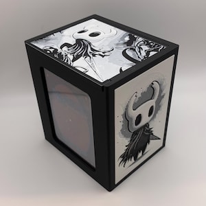 Puede incluir: Caja negra para guardar cartas con una ilustración en blanco y negro de un personaje con una máscara con cuernos. La caja tiene un marco negro y una ventana transparente.