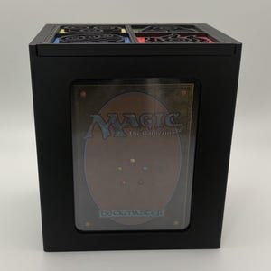May include: Black box with a decorative top and a clear window displaying a Magic: The Gathering card. The card has a brown and gold design with the text "MAGIC The Gathering" and "Deckmaster". The top of the box has a decorative design with red, yellow, blue, and green accents.