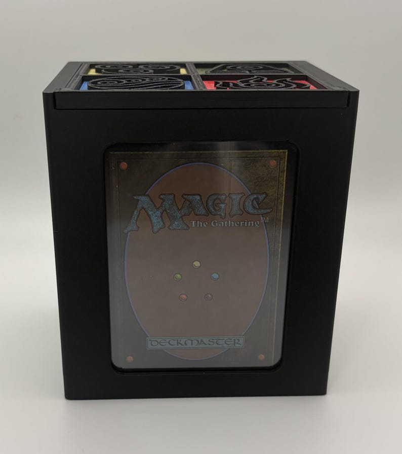 May include: Black box with a decorative top and a clear window displaying a Magic: The Gathering card. The card has a brown and gold design with the text "MAGIC The Gathering" and "Deckmaster". The top of the box has a decorative design with red, yellow, blue, and green accents.