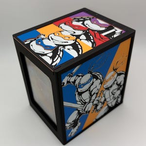 May include: A black rectangular box featuring Teenage Mutant Ninja Turtles graphics. The top panel displays four turtle faces in blue, orange, red, and purple masks. The side panel shows two turtles in action poses against blue and orange backgrounds.