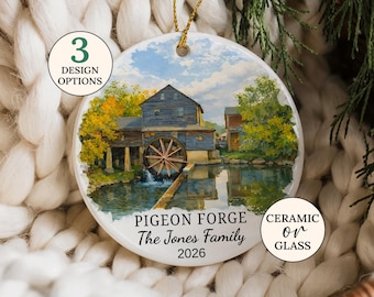 May include: A round ornament featuring a printed illustration of a blue mill with a water wheel by a pond. The text reads PIGEON FORGE, The Jones Family, and 2026. Two circular badges state 3 DESIGN OPTIONS and CERAMIC or GLASS. A gold string is attached.