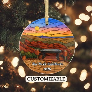 May include: A round ceramic ornament featuring a stained-glass style illustration of the Red Rocks Amphitheatre at sunset. The design includes red rock formations, a stage, and a yellow sun. Text reads Red Rocks Amphitheatre and 2026.