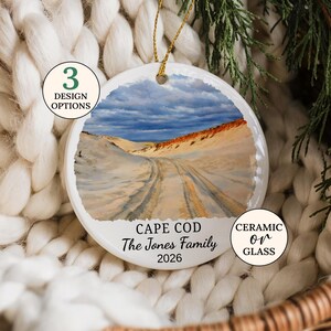 May include: A round white ornament featuring a printed landscape of sand dunes under a cloudy sky, with the text "CAPE COD", "The Jones Family", and "2026" printed below the image. The ornament includes a gold hanging string at the top.