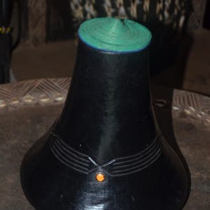 May include: A black conical vessel with a smooth finish, featuring a vibrant green circular lid at the top. The body is decorated with three horizontal embossed lines on each side that converge at a small, centered, round orange accent bead.