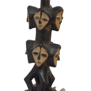 May include: A dark brown wooden sculpture with multiple faces and light tan features. The sculpture has a cylindrical shape with faces arranged around the central column. The base of the sculpture rests on several small stones.