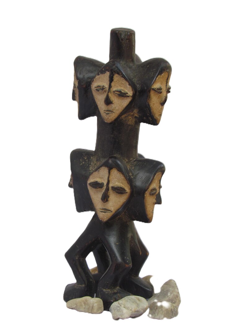 May include: A dark brown wooden sculpture with multiple faces and light tan features. The sculpture has a cylindrical shape with faces arranged around the central column. The base of the sculpture rests on several small stones.