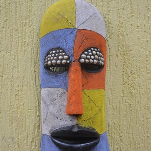 May include: A decorative mask with a face design, made from colourful beads. The mask is divided into sections of yellow, blue, orange, and white. The eyes are filled with small, round objects, and the nose and lips are orange and black.