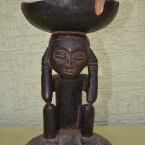 May include: A dark brown hand-carved wooden sculpture of a seated figure with hands raised to the sides of the head, supporting a shallow bowl on top. The wood shows a smooth, polished finish with a small light-colored chip on the bowl's rim.
