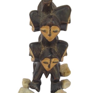 May include: A dark brown wooden sculpture with multiple faces and closed eyes. The sculpture has a stacked design, with a central figure and additional faces around it. Small stones are placed around the base.