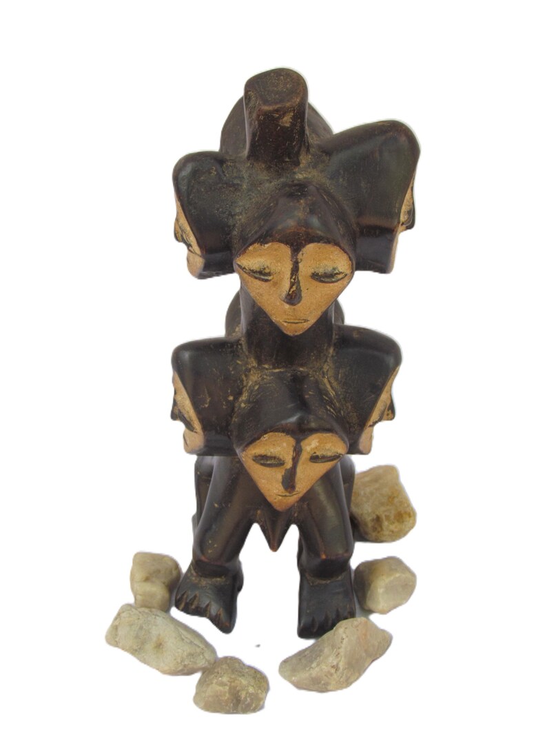 May include: A dark brown wooden sculpture with multiple faces and closed eyes. The sculpture has a stacked design, with a central figure and additional faces around it. Small stones are placed around the base.