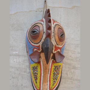 May include: A wooden bird mask featuring a central crest, a pointed beak, and large circular eye sockets. The surface is decorated with intricate patterns of small red, blue, yellow, and white beads, with smooth brown wood accents on the eyes and beak.