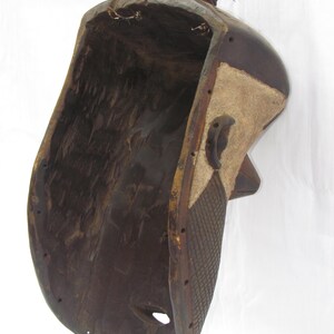 May include: A dark brown, carved wooden mask with a rounded top and a hollowed-out interior. The mask features a textured, patterned side panel with a lighter-coloured section and a small, rounded knob at the top. A hole is visible near the bottom.