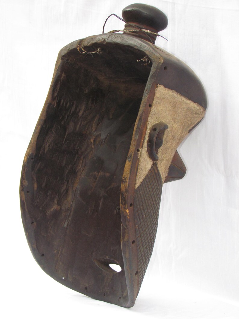 May include: A dark brown, carved wooden mask with a rounded top and a hollowed-out interior. The mask features a textured, patterned side panel with a lighter-coloured section and a small, rounded knob at the top. A hole is visible near the bottom.