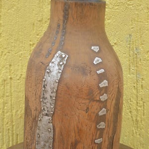 May include: A cylindrical wooden vessel with a natural light brown grain, featuring a vertical metallic strip on the left side and a series of small, individual metallic rectangular inlays arranged in a curved line on the right side.