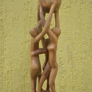May include: A light brown wooden sculpture depicting three stylised figures standing in a circle, their arms extended upwards to collectively support a large, smooth, spherical object held high above their heads on a circular base.
