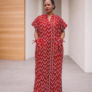 May include: A floor-length red kaftan dress featuring a repeating white geometric diamond pattern, a V-neckline, short sleeves, and two large front pockets with side tie details. The garment has a loose, straight silhouette and reaches the ankles.