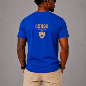 May include: A bright blue short-sleeved t-shirt featuring the gold text CONGO LES LÉOPARDS and a gold leopard head graphic with a star above it on the back. The shirt is paired with light tan trousers, creating a simple and casual appearance.