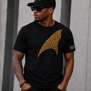 May include: A black short-sleeved t-shirt featuring a large, curved, leopard-print graphic on the chest and a small flag patch on the left sleeve. The outfit includes a matching black baseball cap with a smaller leopard-print logo on the front.