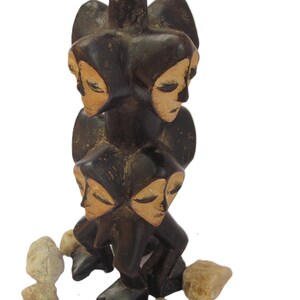 May include: A dark brown wooden sculpture with four faces, each with closed eyes. The sculpture is encircled by small, light-coloured stones. The faces have a light tan colour.