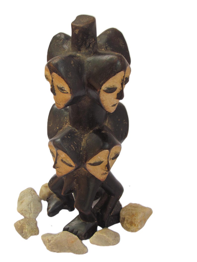 May include: A dark brown wooden sculpture with four faces, each with closed eyes. The sculpture is encircled by small, light-coloured stones. The faces have a light tan colour.