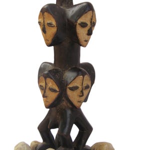 May include: A dark brown wooden sculpture with four carved faces, each with light tan features. The sculpture is set on a base of small stones. The faces are arranged in two pairs, one above the other.