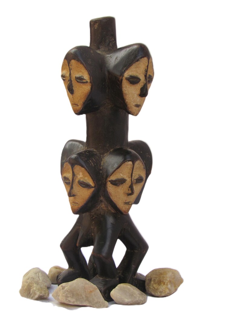 May include: A dark brown wooden sculpture with four carved faces, each with light tan features. The sculpture is set on a base of small stones. The faces are arranged in two pairs, one above the other.