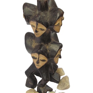 May include: A dark brown wooden sculpture with multiple faces, each with closed eyes. The sculpture has a stacked design, with each face appearing hooded. Small, light-coloured stones are at the base of the sculpture.