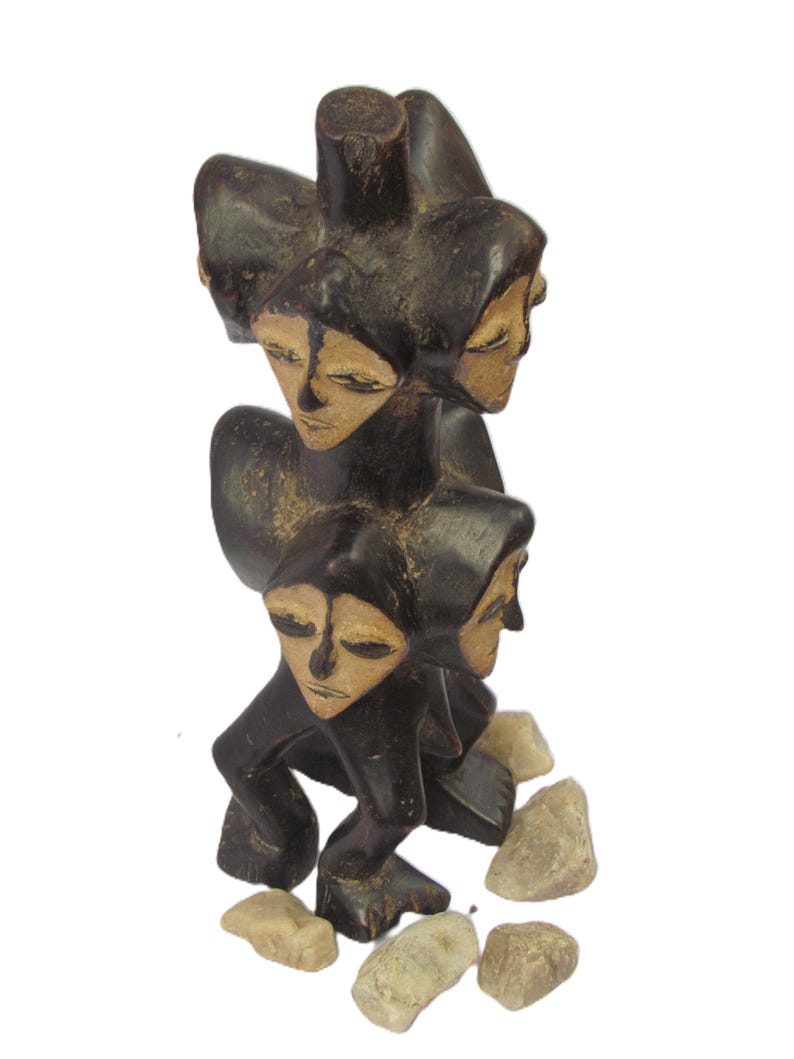 May include: A dark brown wooden sculpture with multiple faces, each with closed eyes. The sculpture has a stacked design, with each face appearing hooded. Small, light-coloured stones are at the base of the sculpture.