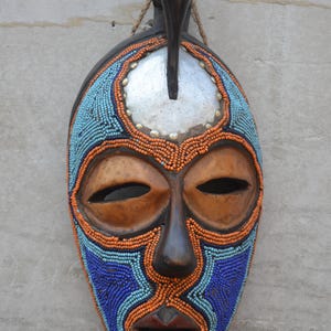 May include: A decorative wall mask featuring a carved wooden face with large almond-shaped eyes and an open mouth. The surface is adorned with intricate patterns of blue, light blue, and orange beads, a silver-toned forehead plate, and a bird figure.