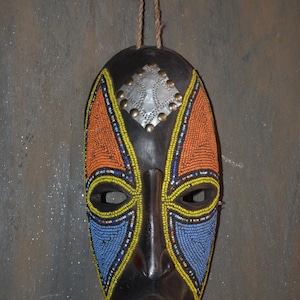 May include: A dark brown wooden mask featuring a central silver-toned metal plate at the forehead, accented with small studs. The mask is decorated with intricate patterns of orange, yellow, and light blue beads around the eyes and cheeks.