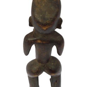 以下が含まれることがあります： 背面から見た、濃い茶色の彫刻が施された木製の人物像。丸い頭、小さな耳、外側に伸びた腕があります。脚は太く、足は離れています。