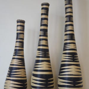 May include: Three tall, slender floor vases of varying heights, each featuring a woven construction with a repeating geometric pattern in alternating bands of black and cream. The vases have a narrow neck and a wider, rounded base design.