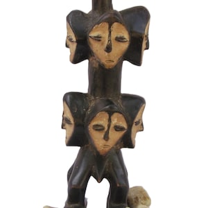 May include: A dark brown wooden sculpture with multiple carved faces, each with closed eyes. The sculpture is vertically designed and rests on light-coloured stones. The faces are a light tan colour.