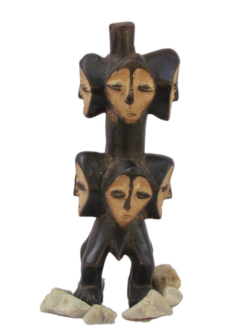 May include: A dark brown wooden sculpture with multiple carved faces, each with closed eyes. The sculpture is vertically designed and rests on light-coloured stones. The faces are a light tan colour.