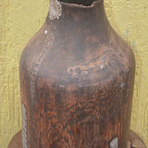 May include: A dark brown wooden vessel with a cylindrical body and a narrow neck. The exterior surface features visible wood grain and is decorated with small, flat, metallic-colored rectangular inlays arranged in vertical and horizontal patterns.